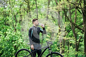 Young man in track-suit with bicycle drinking water at summer park