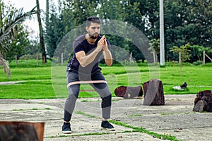 young man training with elastic bands, doing squat leg exercise outdoors in a park