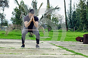 young man training with elastic bands, doing squat leg exercise outdoors in a park