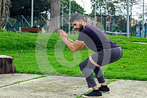 young man training with elastic bands, doing squat leg exercise outdoors in a park