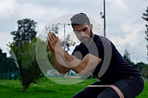 young man training with elastic bands, doing squat leg exercise outdoors in a park