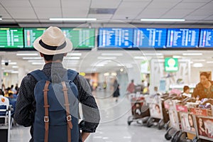 Young man traveler with hat checking flight time, Asian passenger looking to information board in international airport terminal.