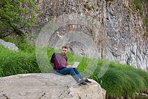 Young man traveller with backpack sits on big flat rock in picturesque place near mountain working on laptop in summer