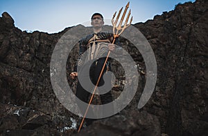 A young man with a trident in his hands against the background of rocks.