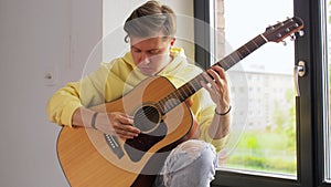 Young man tuning guitar sitting on windowsill