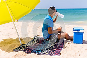 Young man under green solar umbrella drink water from cooler on beach