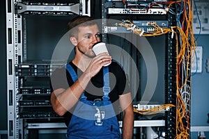 Young man in uniform drinking coffee against internet equipment and wires in server room