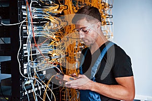 Young man in uniform with measuring device works with internet equipment and wires in server room