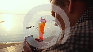 Young man uses cellphone and drinking cocktail on terrace during sunset with sea view. slow motion. 1920x1080