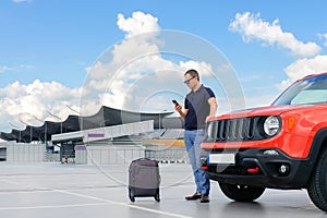 A young man uses a smartphone while standing near a car in an open parking lot. Road lifestyle concept