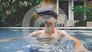 Young man uses virtual reality glasses while swimming in the pool. Guy getting experience in using VR-headset at summer