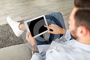 Young man using digital tablet with blank black screen, browsing website or showing new app, sitting on sofa at home