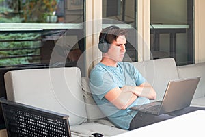 Young man using headphones and laptop while sitting outside on an outdoor couch