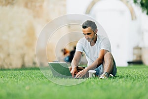 Young man using his laptop on the grass in summer city park