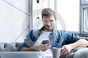 Young man using his smartphone for online banking - sitting on sofa with laptop on leap.