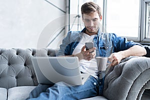 Young man using his smartphone for online banking - sitting on sofa with laptop on leap. photo