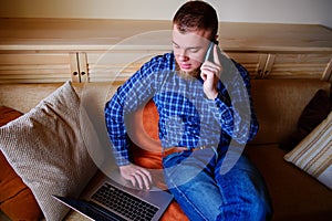 Young man using his smartphone for online banking - sitting on sofa with laptop on leap