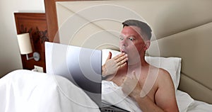 Young man using laptop on bed covers mouth with hand in surprise