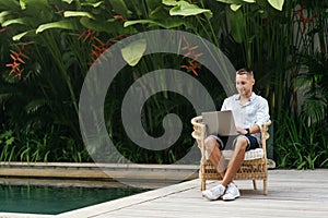 Young man using a laptop computer in a garden with a swimming pool.