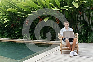 Young man using a laptop computer in a garden with a swimming pool.