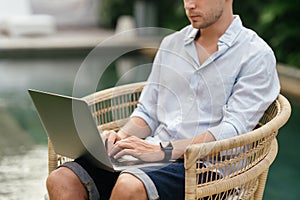 Young man using a laptop computer in a garden with a swimming pool.