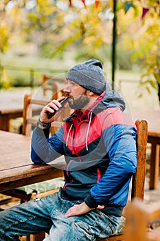 A young man using a mobile phone is sitting outdoors in a cafe or restaurant on an autumn day