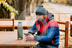 A young man using a mobile phone is sitting outdoors in a cafe or restaurant on an autumn day, drinking a hot drink