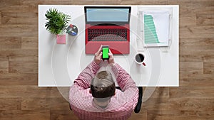 Young man using phone with chroma key, topshot, sitting behind desk with laptop and coffee