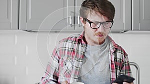 Young man using phone and drinking coffee while sandng in the kitchen