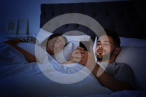 Young man using smartphone while his girlfriend sleeping in bed