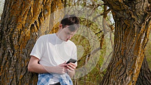 Young man using a smartphone, standing near a large tree in the countryside