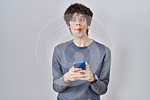 Young man using smartphone typing a message making fish face with mouth and squinting eyes, crazy and comical