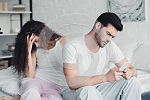 young man using smartphone while upset woman sitting behind on bed relationship