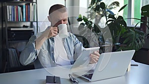 Young man using tablet and drinking coffee while sitting at table in office.
