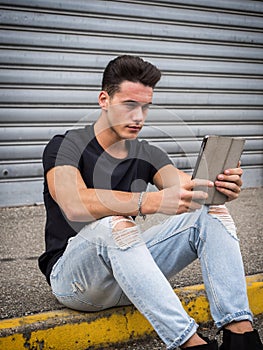 Young man using tablet PC sitting on street curb