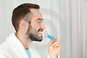 Young man using teeth whitening device on light background.