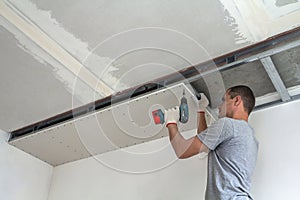 Young man in usual clothing and work gloves fixing drywall suspended ceiling to metal frame using electrical screwdriver on