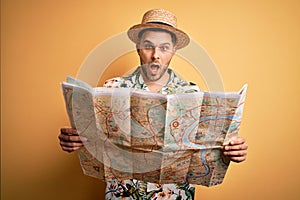 Young man on vacation wearing summer hat holding and looking at tourist map over yellow background scared in shock with a surprise