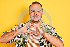 Young man on vacation wearing summer shirt striped t-shirt over isolated yellow background smiling in love showing heart symbol