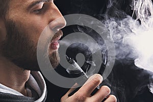 Young man vaping e-cigarette with smoke on black