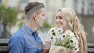 Young man waiting for girlfriend on bench, giving her flowers, happy couple