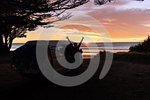 Young man waking up at sunrise by the beach next to his camper van. Van life concept