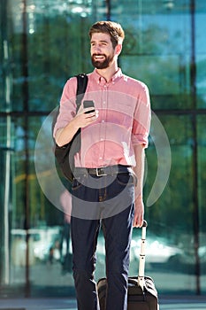 Young man walking in city with cellphone and suitcase