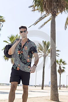 Young man walking on city wearing casual clothes and sunglasses with shorts flowered shirt holding travel bag