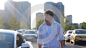 Young man walking near his car on the parking in the city. Buildings on the background. Transport and lifestyle concept