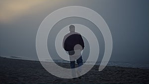 Young man walking on sandy beach in morning fog. Lonely guy looking sea horizon