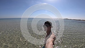 Young man walking with selfie stick on the shallow crystal Sea Water Beach Sand. Concept of relax and happiness.