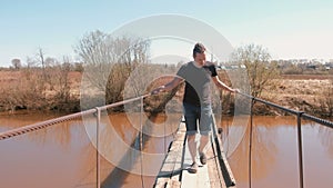 Young man is walking on a suspended wooden bridge over the river.