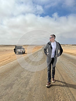 Young man walks on highway, his car at roadside parked in desert. White SUV.