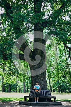 A young man walks in the park. Handsome guy in a T-shirt near a tall tree. Man near a wooden bench in the park